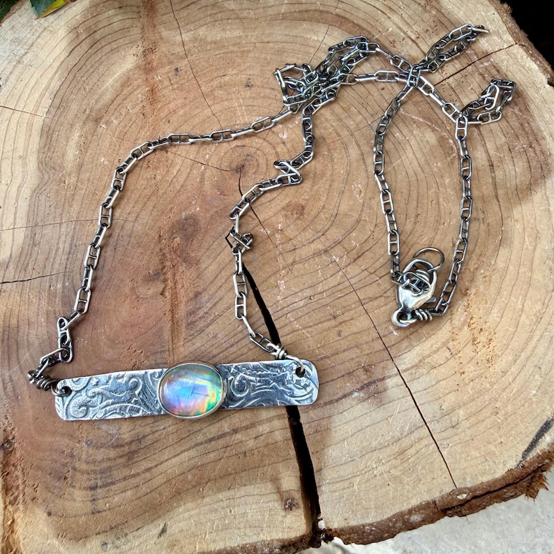 May include: A silver necklace featuring a rectangular metal bar pendant with an embossed floral pattern and a central oval iridescent cabochon stone. The pendant is attached to a silver chain with a lobster claw clasp closure.