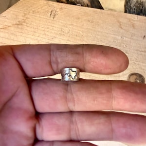 May include: A silver-colored metal ring with a hammered texture and a cutout shape of the state of Texas centered on the band. The ring features a polished finish and a distinct outline of the Texas state border, creating a clear visual contrast against the metallic surface of the band.