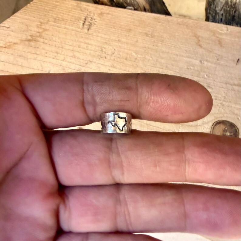 May include: A silver-colored metal ring with a hammered texture and a cutout shape of the state of Texas centered on the band. The ring features a polished finish and a distinct outline of the Texas state border, creating a clear visual contrast against the metallic surface of the band.