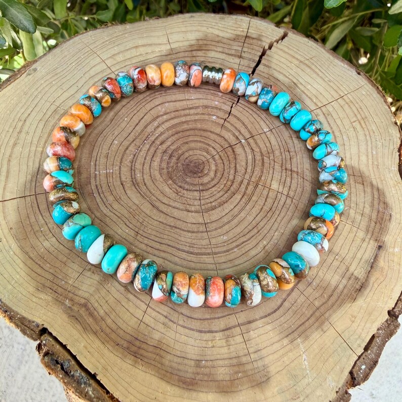 May include: A circular necklace composed of small, flat, disc-shaped beads in shades of turquoise, orange, white, and brown. The beads are arranged in a repeating pattern, interspersed with a single metallic silver-toned cylindrical bead.
