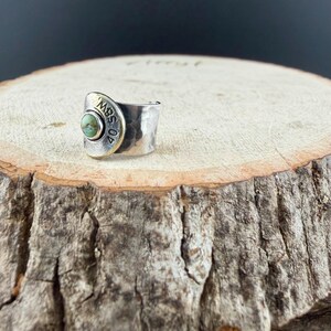 May include: A silver ring with a hammered texture and a gold-coloured accent. The ring features a round, light green stone and the text "40 SBW 02" on the gold accent. The ring is set on a wooden surface.