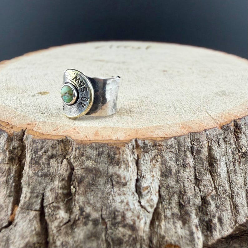 May include: A silver ring with a hammered texture and a gold-coloured accent. The ring features a round, light green stone and the text "40 SBW 02" on the gold accent. The ring is set on a wooden surface.