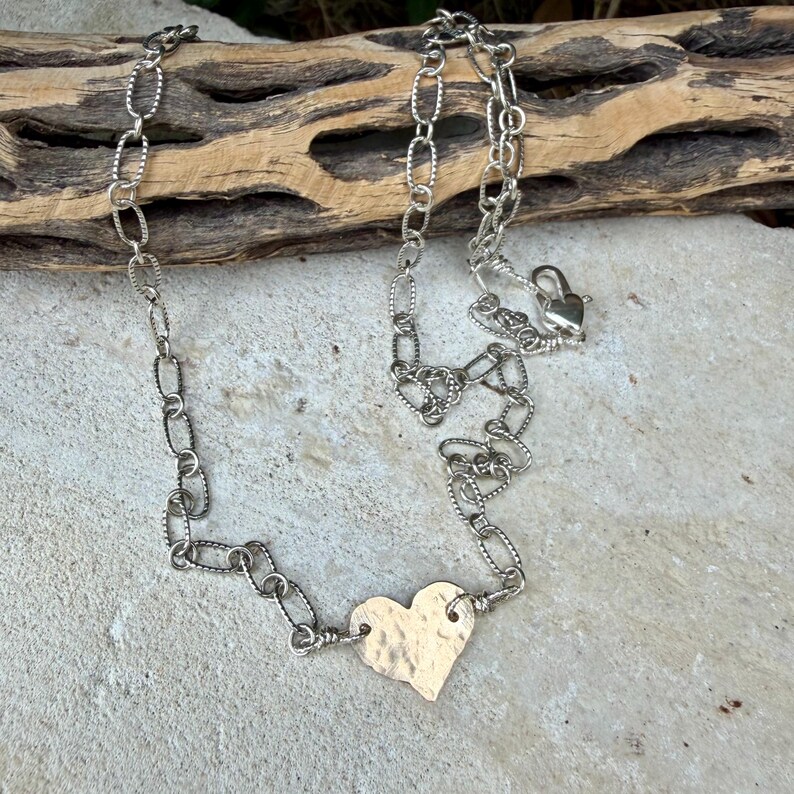 May include: A silver-toned necklace featuring a chain of textured oval links and a single, central heart-shaped pendant with a hammered finish. The necklace includes a lobster claw clasp closure at the end of the chain for secure fastening.