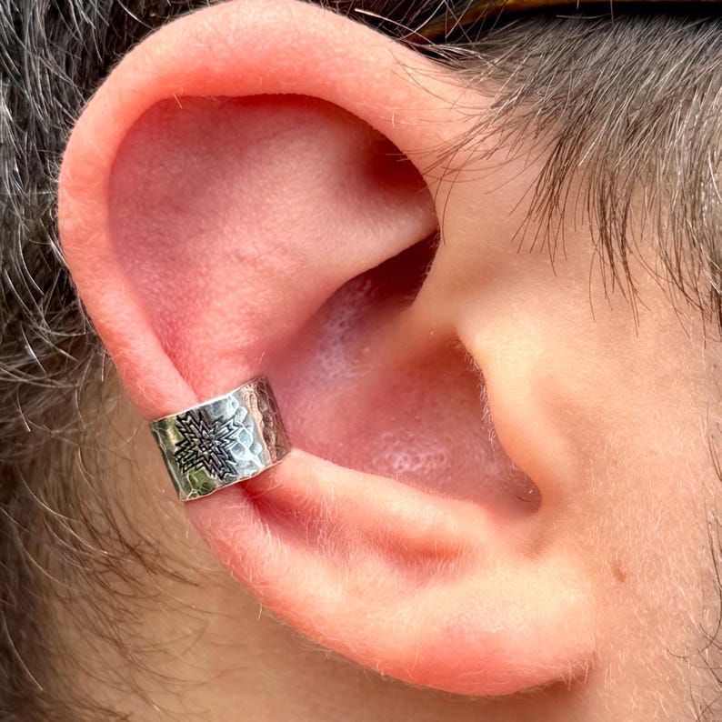 Puede incluir: Un pendiente tipo ear cuff de plata con textura martillada y un patr&oacute;n de cruz geom&eacute;trica grabado en oscuro. La banda de metal es ancha y se ajusta al borde exterior del cart&iacute;lago de la oreja, con un acabado pulido y detalles oxidados.