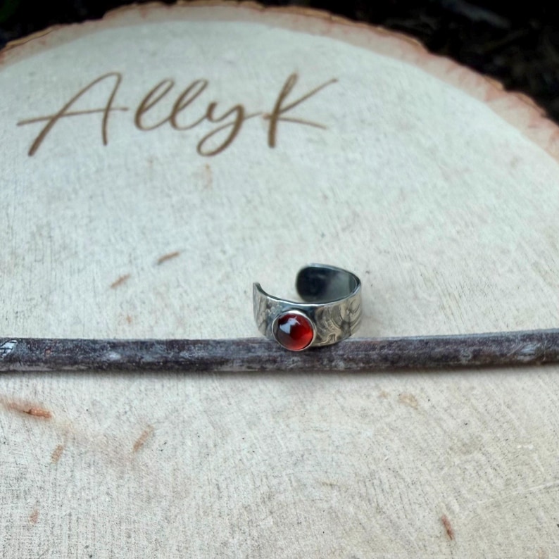 May include: A silver-toned ring with a textured band and a round, red gemstone. The ring is displayed on a wooden surface with the handwritten text "Allyk" in the background.
