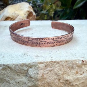 May include: A copper cuff bracelet featuring a repeating geometric pattern of etched diamonds, chevrons, and horizontal lines. The metal has a warm, reddish-brown metallic finish with a polished surface and an open back design for adjustment.