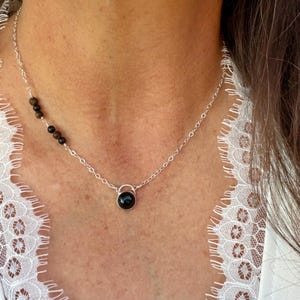 May include: A silver chain necklace featuring a single round black stone pendant at the center and a series of four small, dark brown and black spherical beads arranged asymmetrically along the upper left side of the delicate metal link chain.