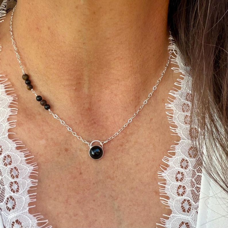 May include: A silver chain necklace featuring a single round black stone pendant at the center and a series of four small, dark brown and black spherical beads arranged asymmetrically along the upper left side of the delicate metal link chain.