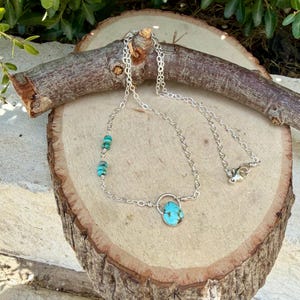 May include: A silver chain necklace featuring a teardrop-shaped turquoise pendant suspended from a small silver circle, with two additional small turquoise beads integrated into the chain on one side and a lobster claw clasp closure.