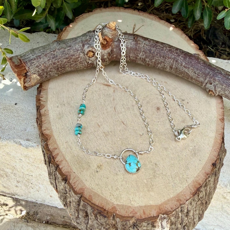 May include: A silver chain necklace featuring a teardrop-shaped turquoise pendant suspended from a small silver circle, with two additional small turquoise beads integrated into the chain on one side and a lobster claw clasp closure.