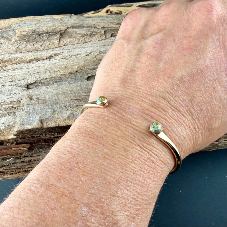 May include: A gold-colored open cuff bracelet with turquoise-colored stone accents. The bracelet curves around the wrist, with a stone at each end. The bracelet is shown on a person's wrist.