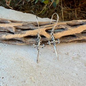 May include: A pair of silver-toned drop earrings featuring a cross-shaped design with ornate, pointed ends and a long, slender vertical spike extending downwards. The earrings are suspended from curved wire hooks and exhibit a polished metallic finish.
