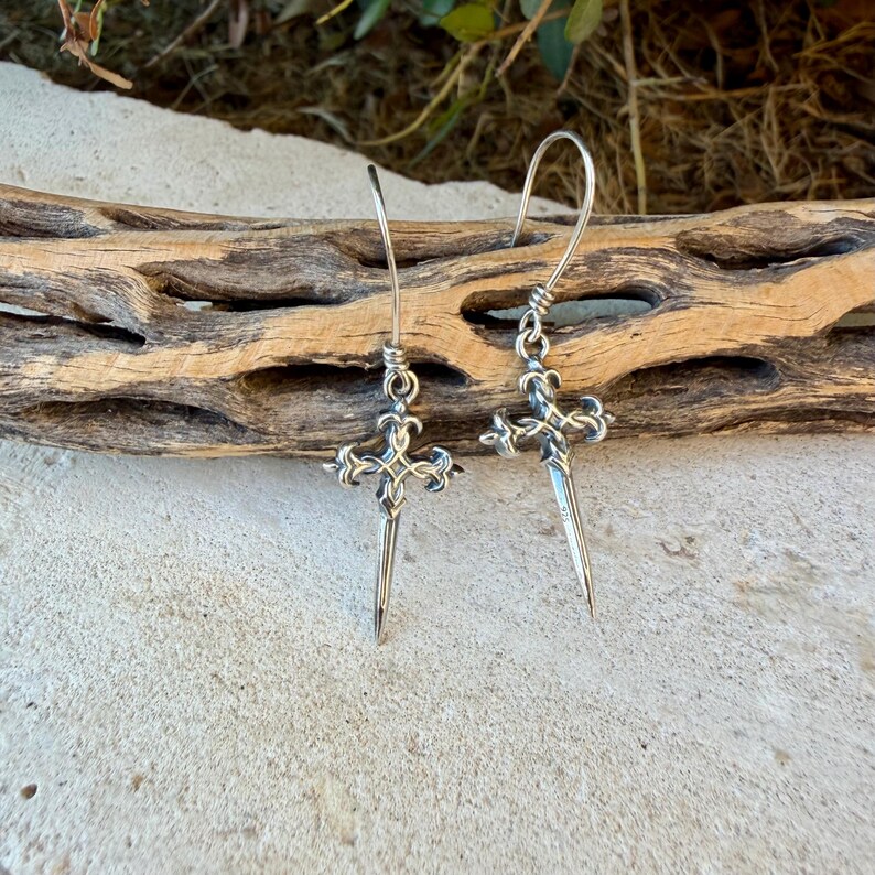 May include: A pair of silver-toned drop earrings featuring a cross-shaped design with ornate, pointed ends and a long, slender vertical spike extending downwards. The earrings are suspended from curved wire hooks and exhibit a polished metallic finish.