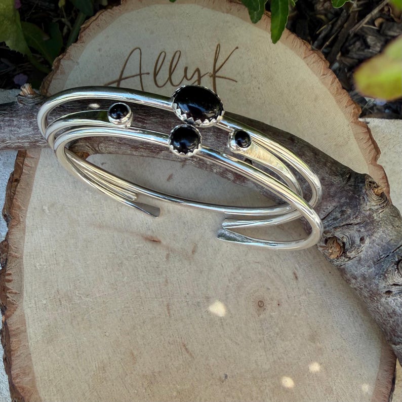 Op de afbeelding: Zilverkleurige manchetarmbanden met zwarte edelsteenaccenten. De armbanden zijn gestapeld, met drie banden zichtbaar. De edelstenen zijn ovaal en rond, gezet in een zilveren bezel. De armbanden worden op een houten oppervlak gepresenteerd.