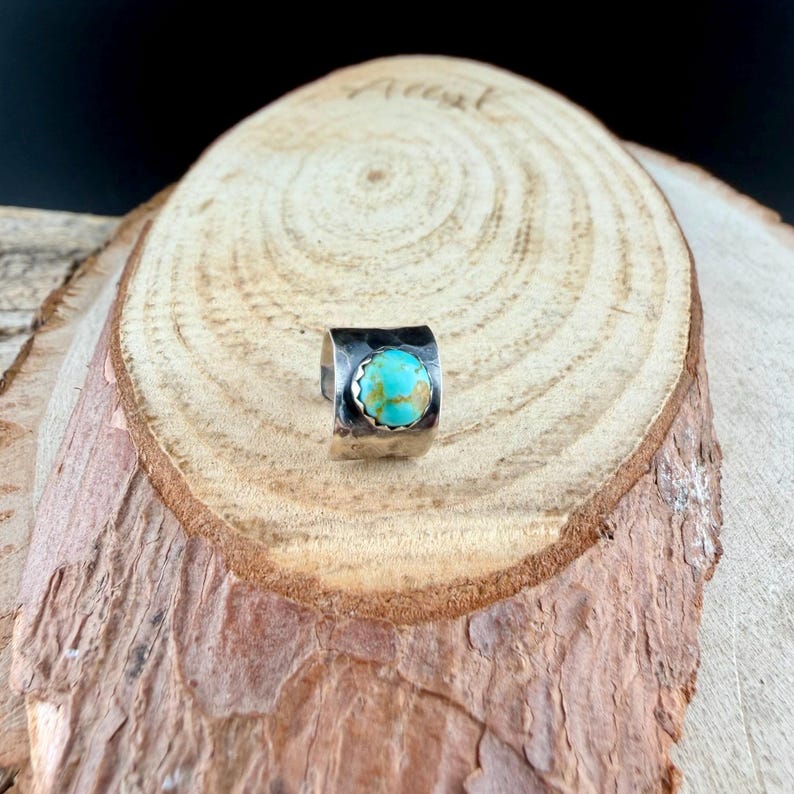 May include: A silver ring with a hammered texture and a round, turquoise stone. The ring is set on a wooden surface with a natural, rustic appearance. The turquoise stone is a vibrant blue-green color.