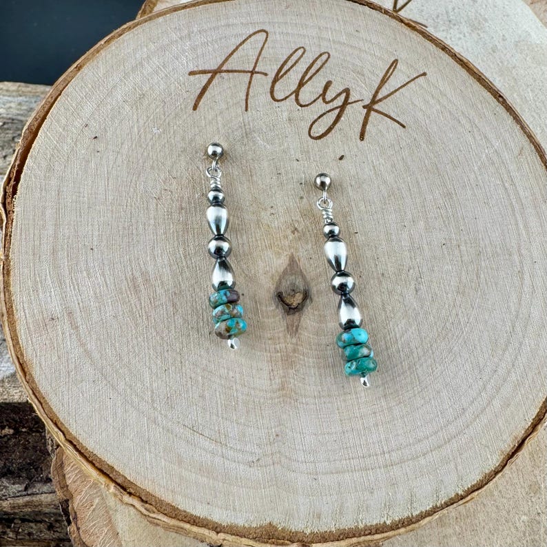 May include: A pair of silver earrings with a post backing. Each earring features a vertical design with alternating silver beads and turquoise stones. The earrings are displayed on a wooden surface with the handwritten text "Allyk".