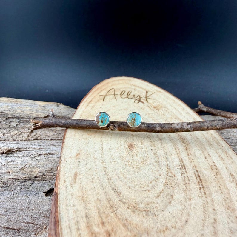May include: A pair of round stud earrings with turquoise-colored centers and silver-colored settings. The earrings are displayed on a small wooden branch, resting on a wooden surface. The name "Allyk" is written on the wood.