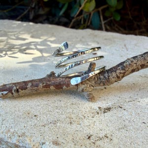 May include: A silver-colored metal ring featuring a spiral design with a hammered, textured finish. The band wraps around itself multiple times, creating a layered appearance with tapered ends. The surface of the metal reflects light, highlighting the indentations created by the hammering process.