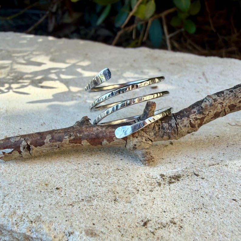 May include: A silver-colored metal ring featuring a spiral design with a hammered, textured finish. The band wraps around itself multiple times, creating a layered appearance with tapered ends. The surface of the metal reflects light, highlighting the indentations created by the hammering process.