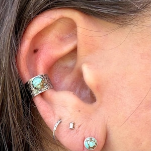 May include: Close-up of an ear adorned with multiple pieces of jewelry. A silver ear cuff with a turquoise stone sits on the upper ear. Additional earrings include a small hoop, a rectangular stud, and a turquoise stud.