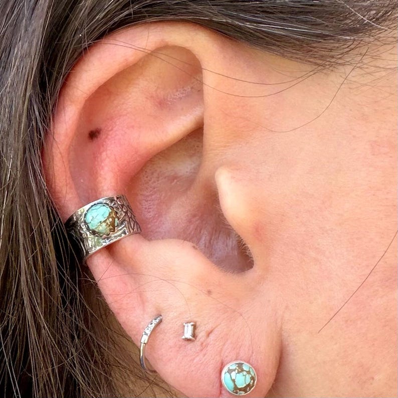 May include: Close-up of an ear adorned with multiple pieces of jewelry. A silver ear cuff with a turquoise stone sits on the upper ear. Additional earrings include a small hoop, a rectangular stud, and a turquoise stud.
