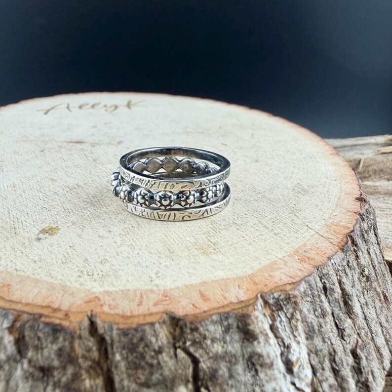 May include: Three silver rings stacked on top of each other. The top ring has a series of small, evenly spaced circles. The middle ring has a floral design. The bottom ring has a series of small, evenly spaced floral designs.