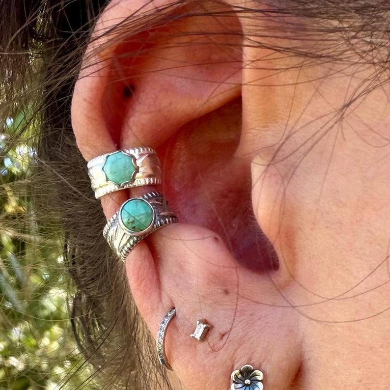 May include: Two silver ear cuffs featuring round turquoise stones with scalloped edges, worn alongside a thin silver hoop, a small rectangular stud, and a silver flower-shaped stud earring on the lower ear lobe.