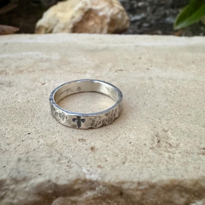 May include: A silver ring with a hammered texture and a small cross design. The ring is set against a neutral background, highlighting its simple yet elegant design. The ring's band is approximately 0.25 inches wide.