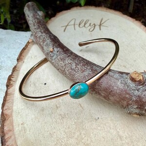 May include: A gold-colored cuff bracelet with a turquoise stone set in the center. The bracelet is resting on a wooden branch and a wooden slice. The name "Ally K" is engraved on the wooden slice.