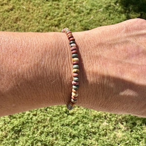 May include: A beaded bracelet with a variety of earth-toned beads, including shades of red, brown, green, and yellow. The bracelet is strung on a flexible cord and is worn on a wrist. The bracelet is a simple, yet elegant piece of jewelry.