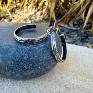 May include: A pair of silver hoop earrings with a textured, braided design. The earrings are displayed on a dark gray stone, with a blurred background of greenery.