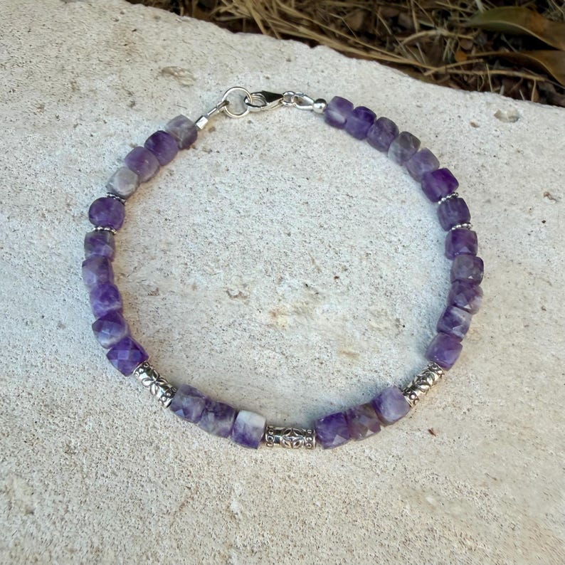 Puede incluir: Una pulsera que presenta cuentas c&uacute;bicas de amatista morada facetada dispuestas en un patr&oacute;n circular. El dise&ntilde;o incluye peque&ntilde;as cuentas espaciadoras de metal plateado y un cierre de mosquet&oacute;n plateado, creando una pieza de joyer&iacute;a sim&eacute;trica.