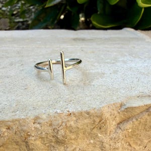 May include: A silver ring with a minimalist design. The ring features two vertical bars, one on each side of the finger, creating a modern and geometric look. The ring is set against a neutral background.