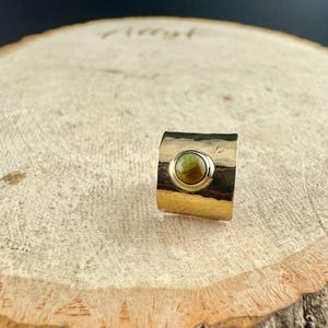 May include: A gold-toned ring with a hammered texture and a circular, olive-green stone set in the center. The ring has a wide band and a simple, elegant design, suitable for everyday wear.