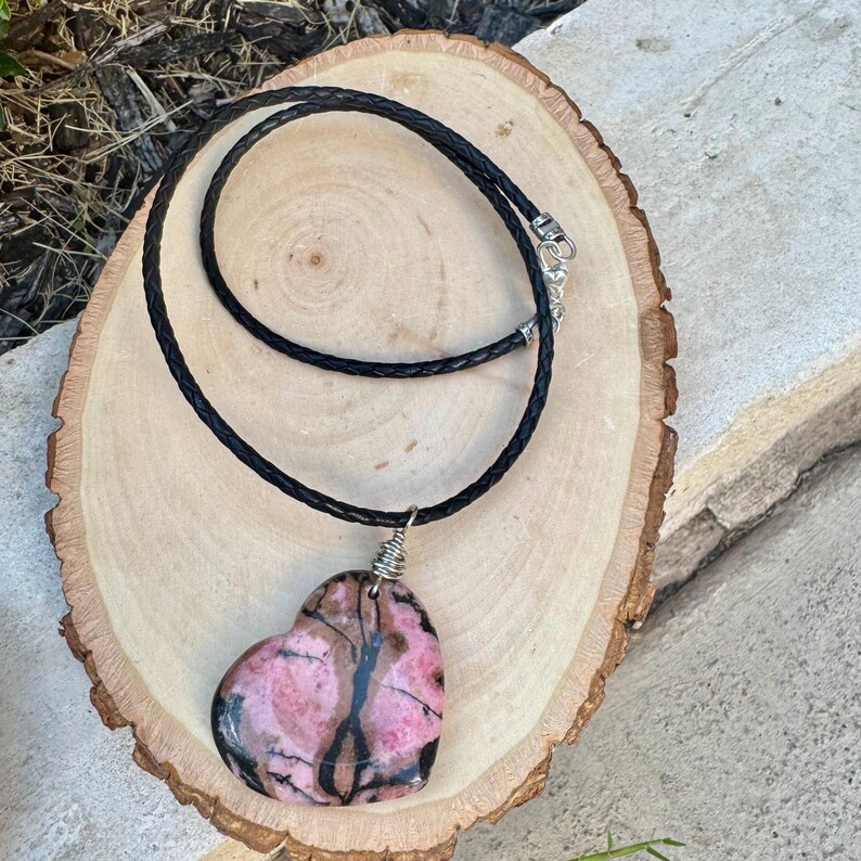 May include: A heart-shaped pendant necklace. The pendant is a pink and black stone, wire-wrapped and suspended from a black braided cord. The necklace is displayed on a light brown wooden surface.