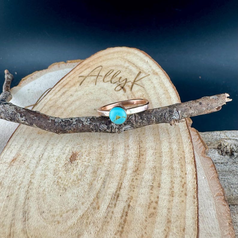 May include: A copper-coloured ring featuring a round, turquoise stone. The ring is presented on a small wooden branch, placed on a wooden slice. The name "Ally K" is etched into the wood. The background is dark.