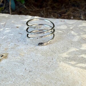 May include: A silver-colored metal ring featuring a multi-coil spiral design. The band has a textured, hammered finish across its surface, with rounded, tapered ends that complete the continuous wrap-around structure of the piece.
