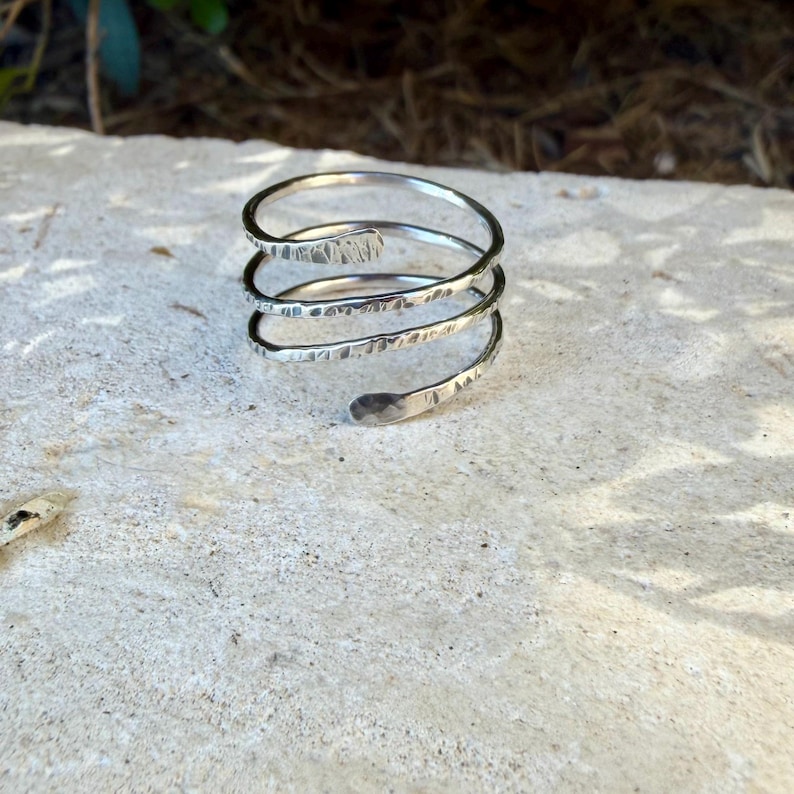 May include: A silver-colored metal ring featuring a multi-coil spiral design. The band has a textured, hammered finish across its surface, with rounded, tapered ends that complete the continuous wrap-around structure of the piece.