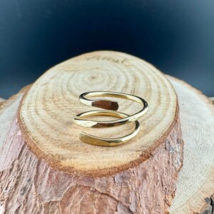May include: A gold-colored, spiral ring with a hammered texture. The ring wraps around the finger with a three-turn design. The jewelry is displayed on a wooden surface.