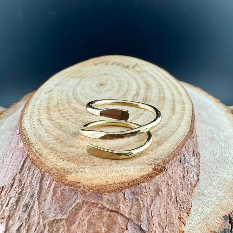 May include: A gold-colored, spiral ring with a hammered texture. The ring wraps around the finger with a three-turn design. The jewelry is displayed on a wooden surface.