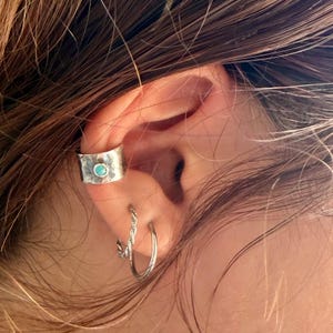 May include: A silver ear cuff with a hammered texture and a small round turquoise stone set in the center, worn alongside a thin twisted silver hoop earring and a plain silver hoop earring on the lower lobe of the ear.