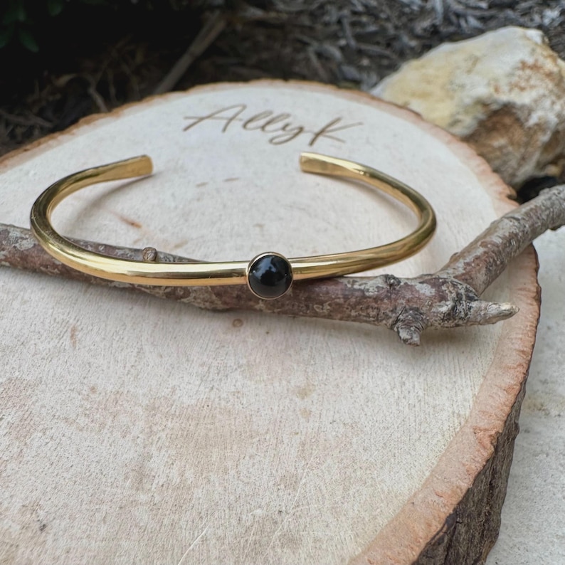 May include: A gold-colored cuff bracelet with a small, round, black stone in the center. The bracelet is displayed on a wooden surface with the name "Ally K" etched into the wood.