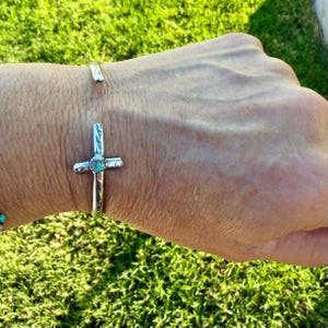 May include: A silver-toned cuff bracelet with a cross pendant featuring a turquoise stone. The cross is centered on the wrist, with a second, smaller silver cuff visible above. The bracelet is displayed against a background of green grass.