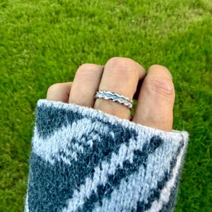 May include: A silver ring featuring a textured, wavy, or scalloped edge design along the band. The ring is worn on a finger, with the band displaying a repeating pattern of small, raised, rounded shapes that create a distinct, undulating profile.