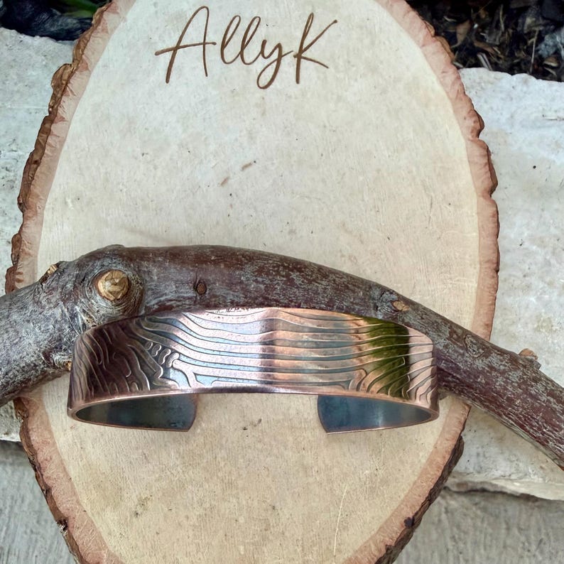 May include: A copper cuff bracelet with an etched design, resting on a wooden slice. The bracelet has a textured pattern and is displayed with a natural, rustic aesthetic. The name "Allyk" is written in cursive on the wood.