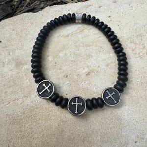 May include: Black beaded bracelet with three silver-toned cross charms. The bracelet features a cylindrical silver bead and is displayed on a light-colored stone surface.