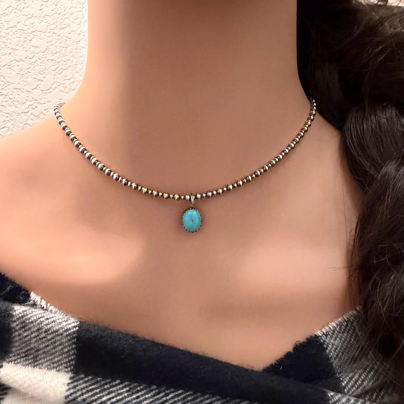 May include: A delicate beaded necklace with a small, round turquoise pendant. The necklace features a mix of silver and gold-toned beads. The pendant is a vibrant turquoise color, adding a pop of color to the design. The necklace is worn around the neck.