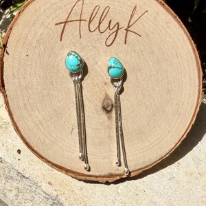 May include: A pair of dangle earrings featuring teardrop-shaped turquoise stones with dark brown matrix patterns. Each earring has two long, thin, hammered silver-tone metal stems extending downward from the stone, ending in small rounded tips.