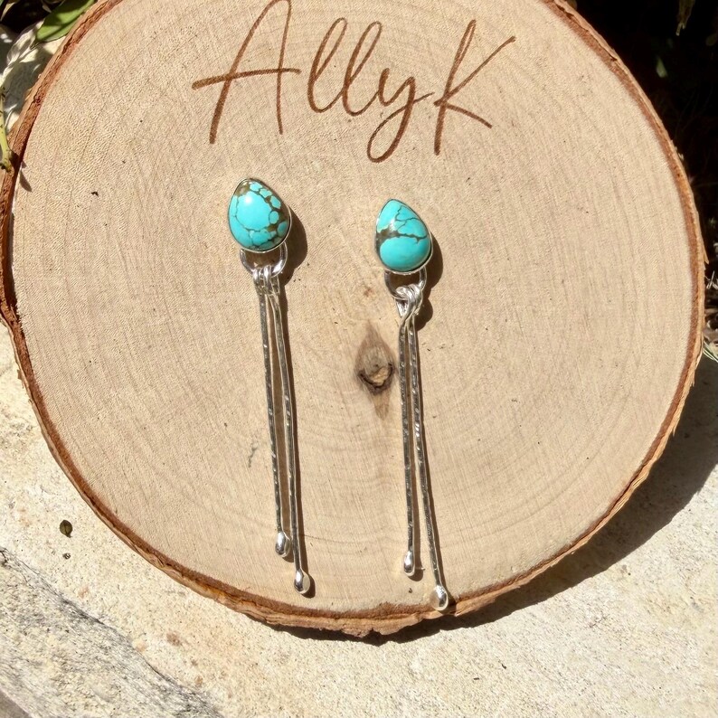May include: A pair of dangle earrings featuring teardrop-shaped turquoise stones with dark brown matrix patterns. Each earring has two long, thin, hammered silver-tone metal stems extending downward from the stone, ending in small rounded tips.