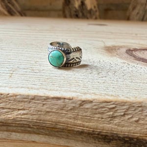 May include: A silver ring featuring a round, polished turquoise stone set in the center. The band is crafted from oxidized silver with a textured surface, displaying etched geometric patterns and a beaded border detail along the edges.